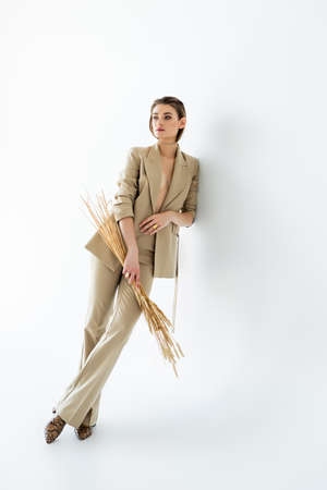full length of young woman in beige formal wear posing while holding wheat on whiteの写真素材
