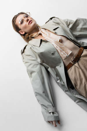 high angle view of trendy woman in glasses, trench coat and scarf lying while posing on whiteの写真素材
