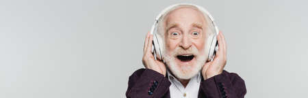 Excited senior man listening music in wireless headphones isolated on gray, bannerの写真素材