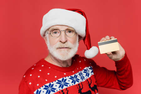 Senior man in sweater and santa hat showing credit card isolated on redの写真素材