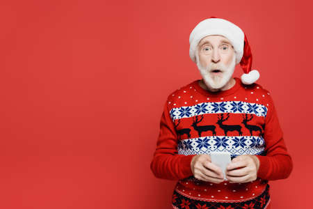 Shocked senior man in santa hat holding smartphone isolated on red