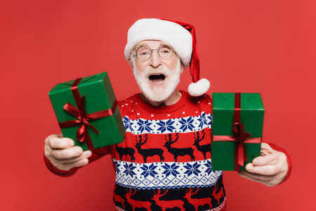 Cheerful elderly man in santa hat holding gift boxes on blurred foreground isolated on red