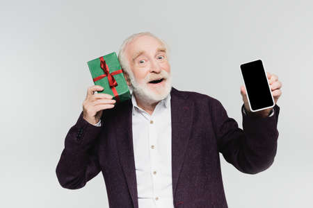 Excited senior man holding smartphone with blank screen and gift isolated on gray