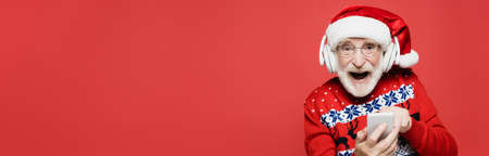 Cheerful senior man in santa hat and headphones using smartphone isolated on red, banner