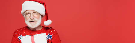 Smiling senior man in santa hat holding present isolated on red, bannerの写真素材