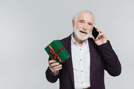 Smiling senior man holding gift while talking on smartphone isolated on gray