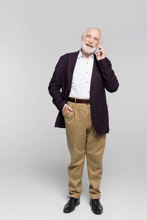 Smiling elderly man with hand in pocket of pants talking on smartphone on gray backgroundの写真素材