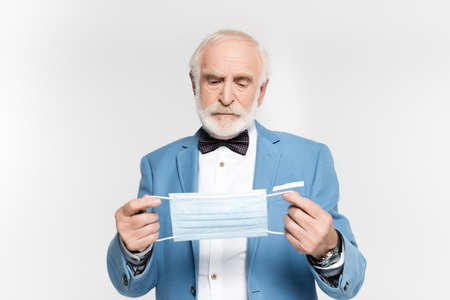 Senior man in jacket and bow tie holding medical mask isolated on gray