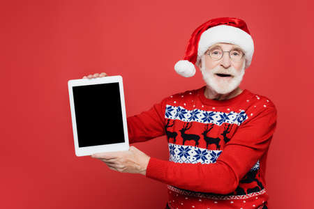 Senior man in santa hat holding digital tablet with blank screen isolated on redの写真素材