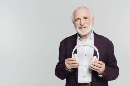 Senior man smiling at camera while holding headphones isolated on gray