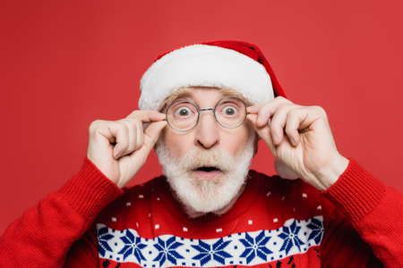 Shocked senior man in santa hat and eyeglasses looking at camera isolated on redの写真素材