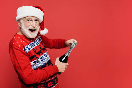 Cheerful senior man in santa hat holding bottle of champagne isolated on redの写真素材