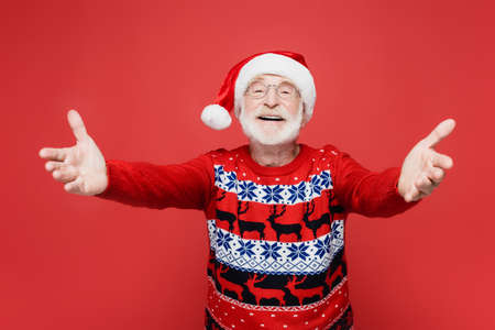 Smiling senior man in santa hat pulling hands at camera on blurred foreground isolated on redの写真素材