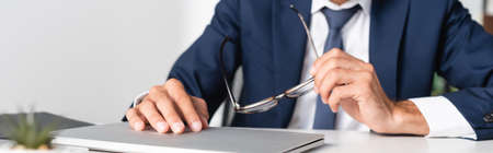 Cropped view of businessman holding eyeglasses near laptop on blurred foreground, bannerの写真素材