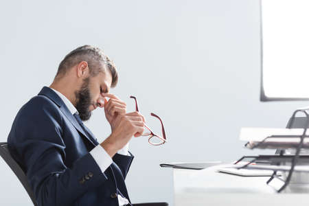 Overworked businessman holding eyeglasses near papers on blurred foreground in office