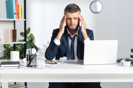 Businessman suffering from headache near gadgets and stationery on blurred foreground in office