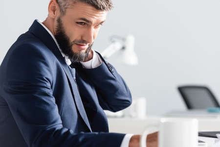 Businessman suffering from neck pain near cup on blurred foreground in office