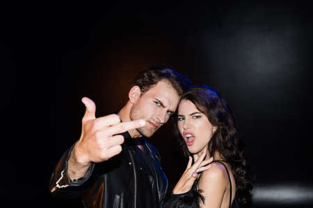 Angry man showing middle finger, standing near brunette woman gesturing on black on blurred...