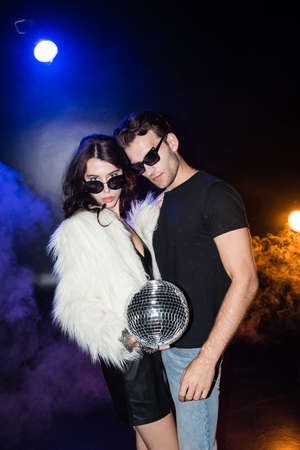 Curly woman holding disco ball, while touching boyfriend shirt and looking at camera with smoke and...