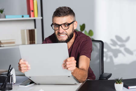 Mad businessman holding laptop near papers folder on blurred foreground in office