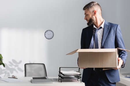Fired businessman holding carton box near office table on blurred background in office