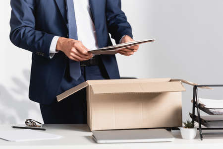 Cropped view of dismissed businessman holding paper folder near laptop and cardboard box on blurred...
