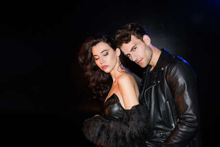 Sensual woman in dress with naked shoulder standing near boyfriend looking at camera on black
