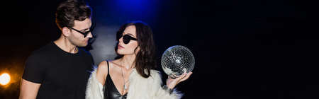 Brunette woman with disco ball looking at man in eyeglasses with backlit on black, banner