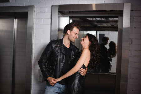 Smiling man in black jacket embracing and holding hand of cheerful curly woman in leather dress near...