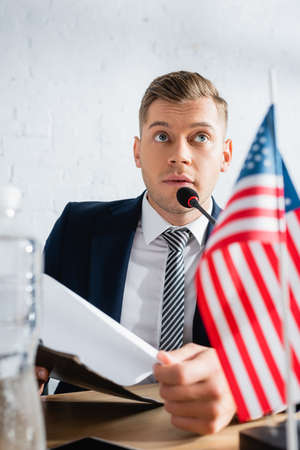 Confident member of political party holding clipboard, while speaking in microphone at table with...