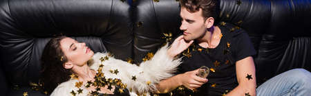 Man looking at passionate woman with glass of champagne lying on sofa, while confetti falling in...