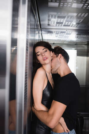 Passionate man in black t-shirt hugging sexy woman with closed eyes, while kissing neck in elevator