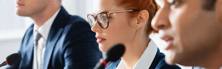 Redhead woman in eyeglasses, sitting near indian man speaking in microphone on blurred foreground,...