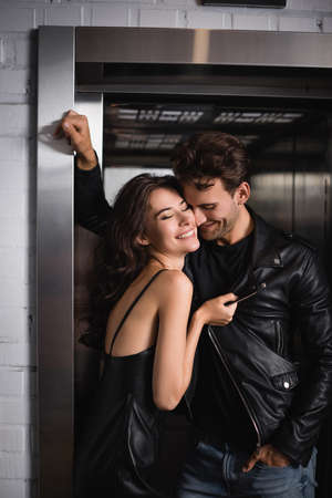 Smiling man with hand in pocket standing near cheerful brunette woman touching jacket, while leaning...