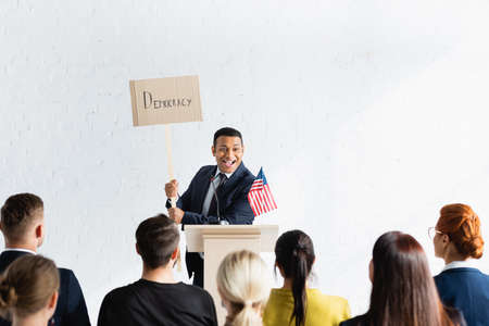 excited indian speaker holding placard with democracy inscription in front of voters in conference...