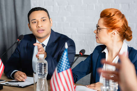 Indian politician gesturing, while talking with colleague, sitting near redhead woman in boardroom...
