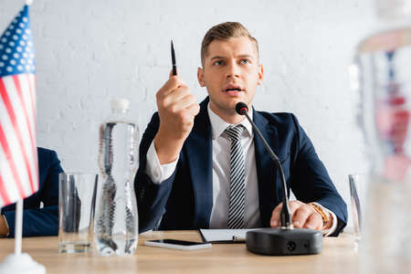 Serious politician gesturing with pen, while speaking in microphone and sitting near colleague on...