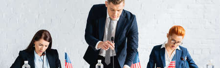Politician leaning forward and touching microphone near female colleagues working in boardroom,...