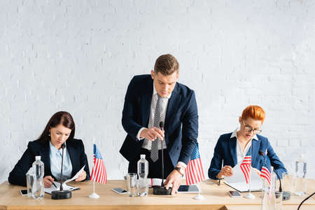 Man in formal wear touching microphone, while leaning forward near female colleagues working in...