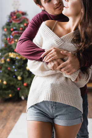 cropped view of happy couple in warm sweaters hugging near decorated christmas tree on blurred backgroundの写真素材