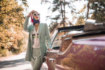 Young elegant woman in sunglasses looking away near car on road on blurred foreground