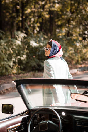 Woman in headscarf and sunglasses standing near auto on blurred foreground on road