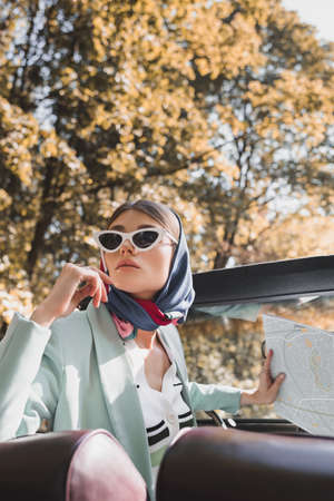 Elegant woman holding map on driver seat of vintage car on blurred foreground