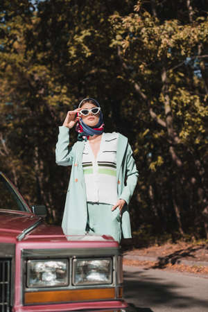 Stylish woman in headscarf touching sunglasses near car on blurred foreground on road