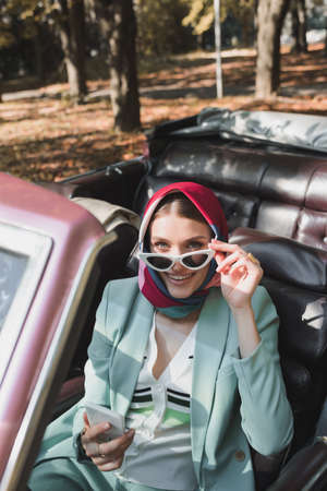 Woman holding sunglasses and smartphone in roofless car on blurred foreground outdoors