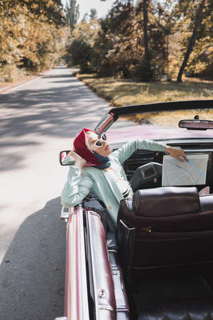 Fashionable woman with map sitting in roofless car on road during trip