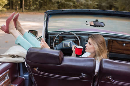 woman putting legs on door while holding cup of coffee in cabriolet