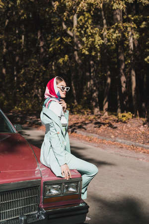 Side view of fashionable woman in sunglasses sitting on car bumper during road trip