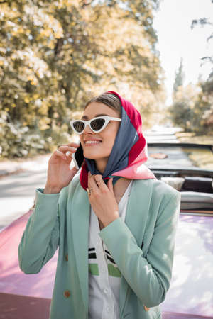Elegant woman smiling while talking on smartphone near vintage car on road