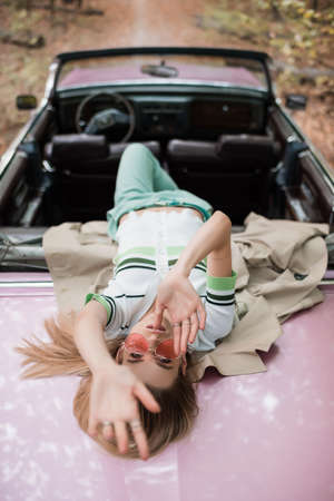 overhead view of young woman looking at camera while lying on cabriolet on blurred backgroundの写真素材